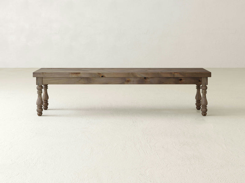 The Annli Bench - Barn Wood by James+James, featuring a dark finish and ornate, turned legs, stands on a plain, light-colored floor against a light-colored, textured wall. The design is simple yet elegant, providing a rustic aesthetic.