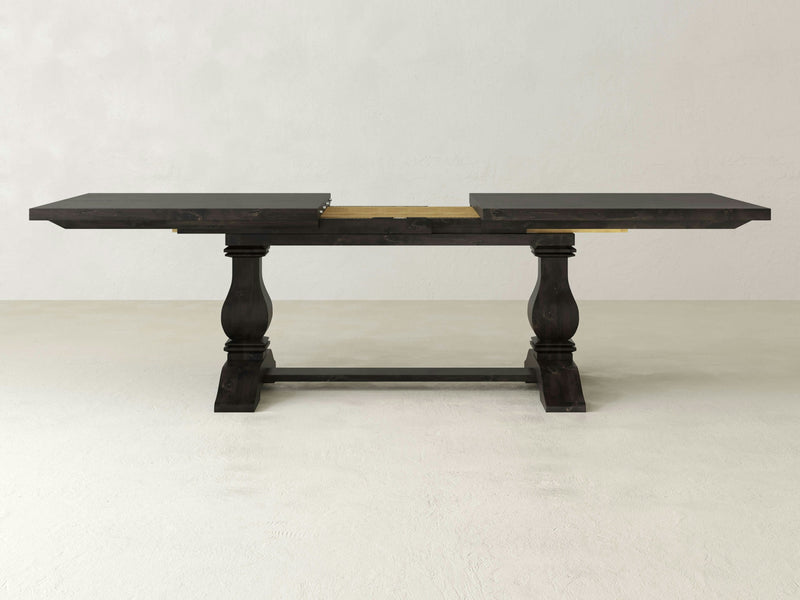 The Heirloom Expandable Dining Table - Charred Ember by James+James is a dark wooden dining table with two pedestal legs featuring intricate carving details. It has an extension mechanism in the center that, when partially extended, reveals a light-colored wood beneath. The background consists of a simple, light grey wall and floor.