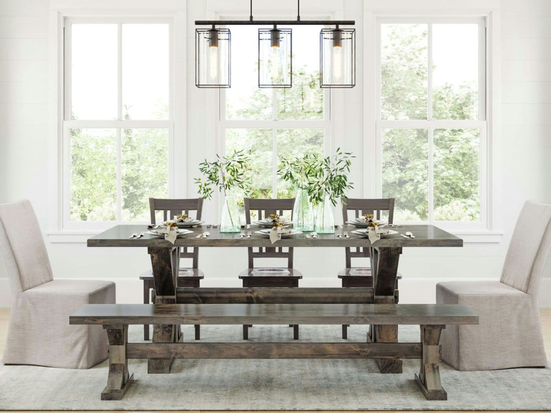 A dining room featuring a rustic wooden table complemented by the Trestle Bench in Deep Grey from James+James, along with matching chairs. The table is adorned with greenery in glass vases and carefully arranged tableware. Two upholstered armchairs grace each end of the table, while a modern, geometric light fixture hangs overhead. Large windows flood the space with natural light.