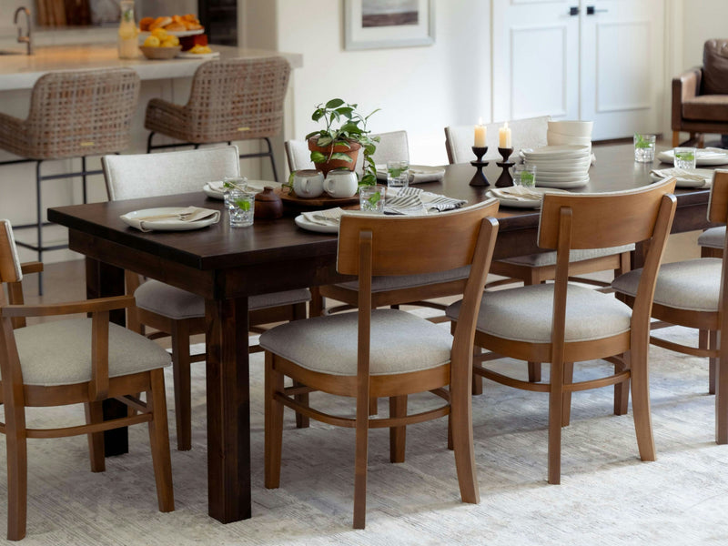 A dining room featuring the Farmhouse Dining Table - Tobacco by James+James elegantly set for a meal. Six wooden chairs with light-colored cushions encircle the table. A potted plant, candles, teacups, and plates are tastefully arranged on the table. In the background, a kitchen island with wicker barstools can be seen.