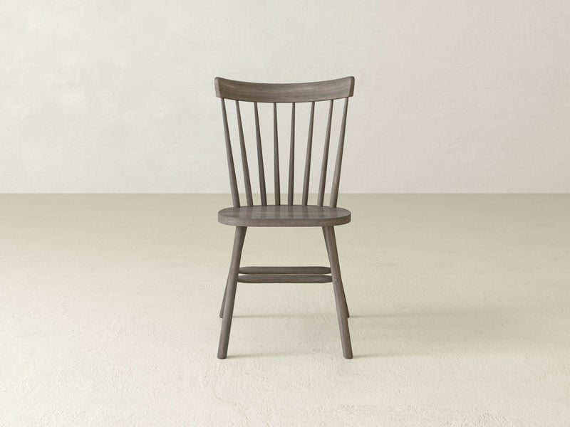 A Rustic Windsor Dining Chair - Barn Wood by James+James, featuring a slightly curved backrest with vertical slats and four tapering legs, is placed in a minimalistic, light-colored room. The chair's finish is a medium-dark tone, complementing its simplistic, classic design.