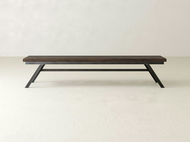 The Fulton Bench - Tobacco by James+James, featuring a modern and minimalist design with a dark brown wooden top and black angular metal legs, is placed against a plain white background. This sleek bench boasts a clean design that's perfect for contemporary settings.