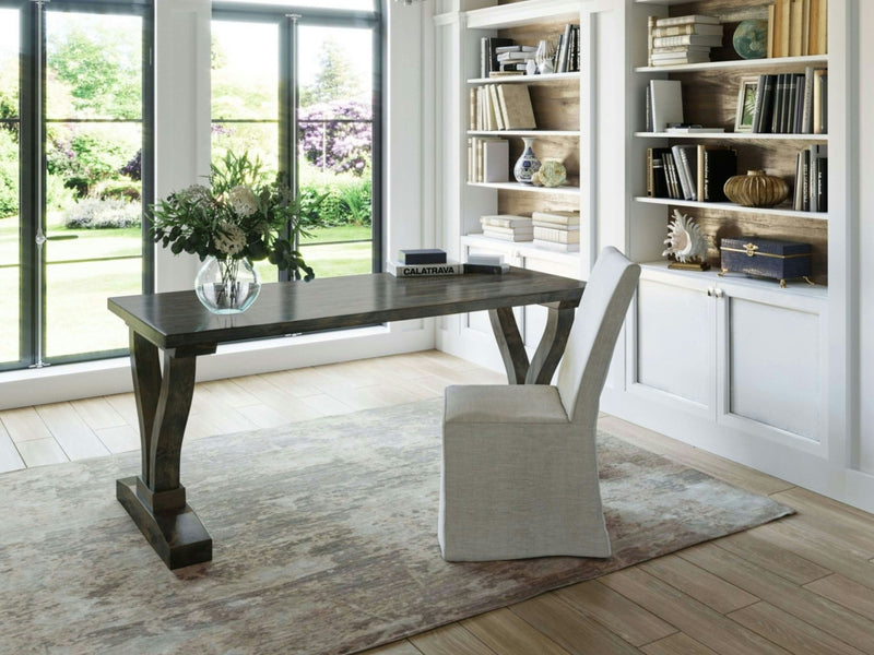 A bright home office features a sleek James+James Vera Desk in deep grey with crossed legs, accompanied by a gray upholstered chair and a vase of flowers on the desk. Behind the desk, built-in white shelves hold books and decorative items. Large windows offer a view of a green, sunlit garden.