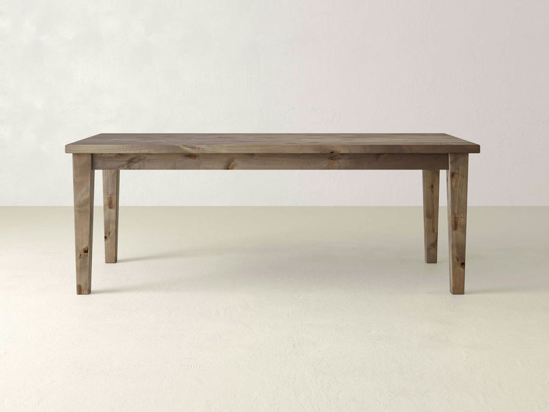 The Ava Parsons Dining Table by James+James showcases a minimalist design with its rectangular shape, four straight legs, and smooth surface. Crafted from barn wood, it features a natural finish that highlights the visible grain and knots. The table is set against a plain, light-colored background, creating a striking contrast.