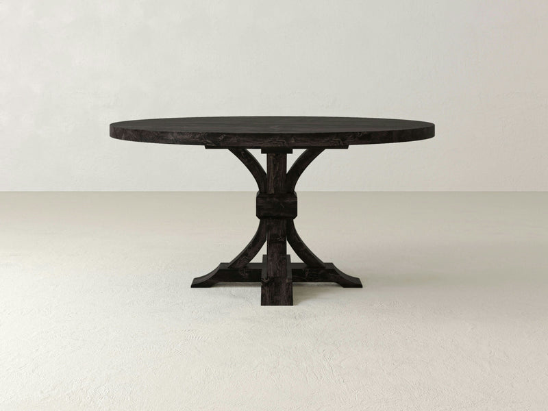 A Violet Round Dining Table by James+James with a dark finish is centered in a minimalist room with light-colored walls and flooring. This solid hardwood pedestal table boasts a thick top and a sturdy, intricately designed base, making it an heirloom-quality centerpiece.