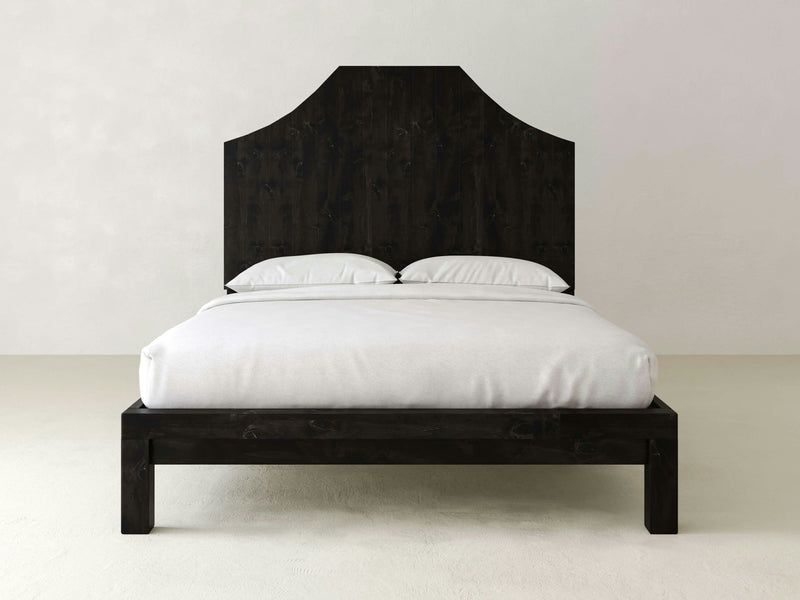 The Megan Bed - Charred Ember by James+James features a minimalist design with a dark, curved headboard. It is adorned with two white pillows and topped with white bedding, set against a neutral, light-colored wall and floor background. The overall design is simple and modern.