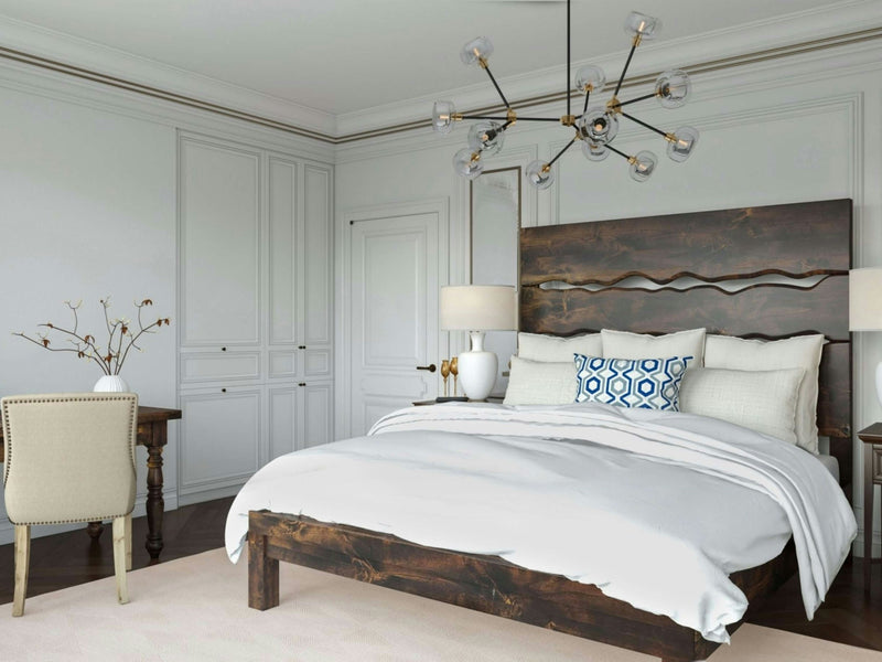 A modern bedroom features the James+James Live Edge Bed in Tobacco with white bedding and a decorative blue and white pillow. Two table lamps with white shades sit on wooden nightstands. A geometric chandelier hangs from the ceiling, and a cushioned chair is placed near a wooden desk.