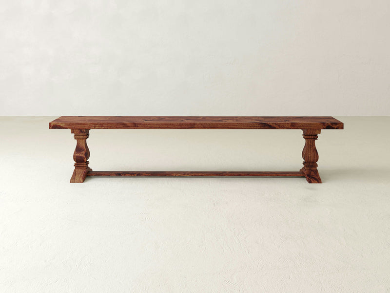 Against a plain beige wall stands the Heirloom Bench - Tuscany from James+James. This wooden bench showcases a rectangular seat with a smooth finish, supported by two intricately carved legs connected by a horizontal bar at the base. The overall design combines minimalist and rustic elements.