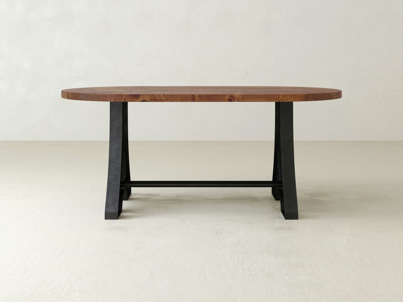 The Wishbone Oval Dining Table - Tuscany by James+James features an elegant wooden tabletop with natural wood grain, providing a rustic touch. It stands on two black metal trestle legs that boast a sleek, modern design. The setting includes a plain, light-colored wall and floor as the background.