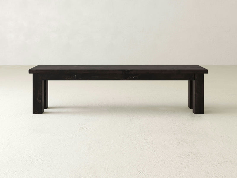 The Farmhouse Bench - Charred Ember by James+James is a minimalist, dark wooden bench with clean lines and no backrest, set against a plain, light-colored background. The bench has a rectangular seat and sturdy legs at each corner, showcasing a simple and modern design.