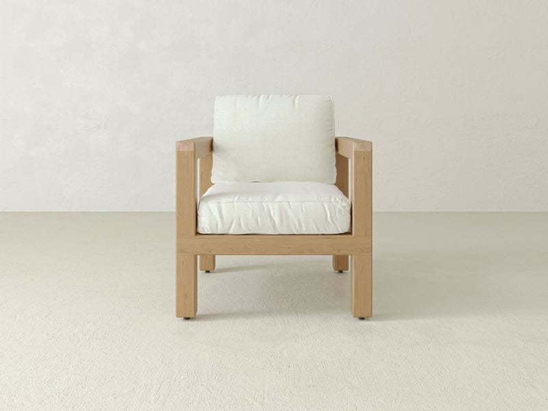 The Aria Outdoor Chair by James+James is a wooden frame armchair featuring a simple, modern design with an off-white cushioned seat and backrest. Positioned against a plain, light-colored wall and floor background, this chair exudes a clean, minimalist aesthetic while offering luxurious comfort for any indoor or outdoor space.