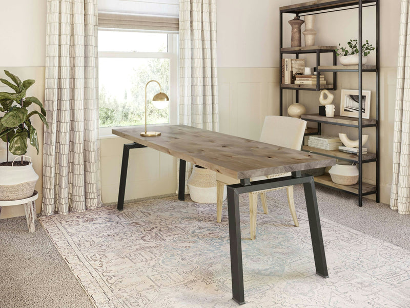 A modern office space featuring the Fulton Desk in Barn Wood by James+James with black metal legs, a beige upholstered chair, and a desk lamp. The room includes a large window with curtains, a potted plant, a patterned rug, and a black metal bookshelf with various decorative items.