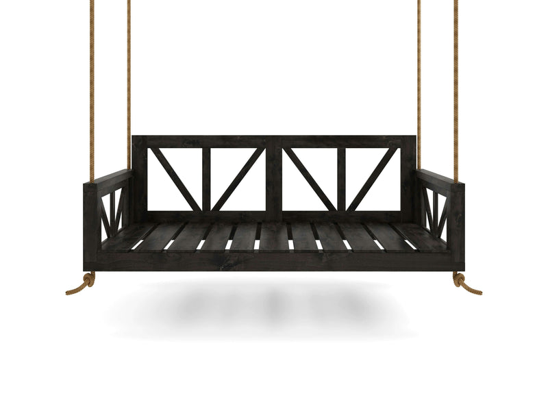 An Avery Porch Swing - Charred Ember by James+James, featuring a dark wooden finish and a simplistic slatted design, hangs from sturdy ropes against a white background. The swing boasts an open backrest with a crisscross pattern and armrests for added comfort. Shadows are cast on the ground beneath it.