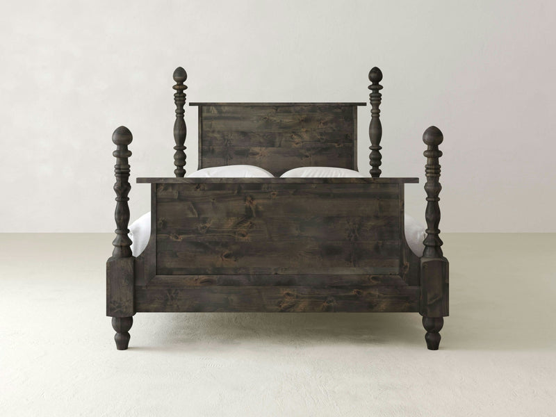 The Catherine Bed by James+James is crafted from hardwood and features four turned posts—two at the headboard and two at the footboard. It stands against a plain, light-colored wall and floor. The wood's dark, slightly distressed finish gives it a rustic appearance with vintage sophistication.