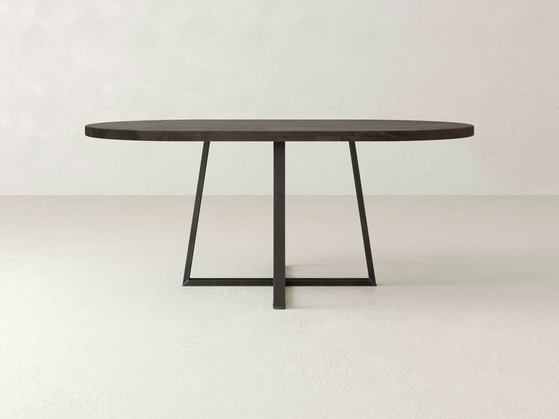 The Watson Oval Dining Table - Deep Grey by James+James stands on a light-colored floor. This minimalist table features an oval wooden top with a deep grey finish and a sleek black metal base. The single support column branches into an X-shaped base at the bottom, creating a contemporary and clean design.