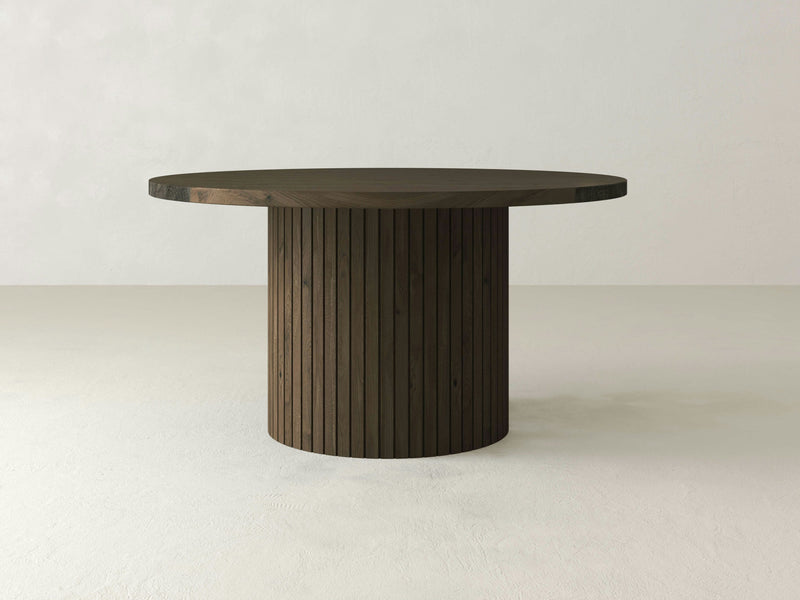 A round wooden Arthur Dining Table - Charred Ember by James+James, featuring a smooth top and a ribbed cylindrical base, set against a plain, light-colored background. The table's design is minimalist and modern with a dark finish.
