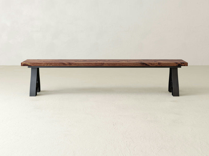 James+James' Wishbone Bench - Tuscany features a minimalist design with a thick, dark brown wood slab seat and sturdy black metal legs. Set against a plain, light-colored background, this modern and simple bench highlights the natural grain and texture of the wood.