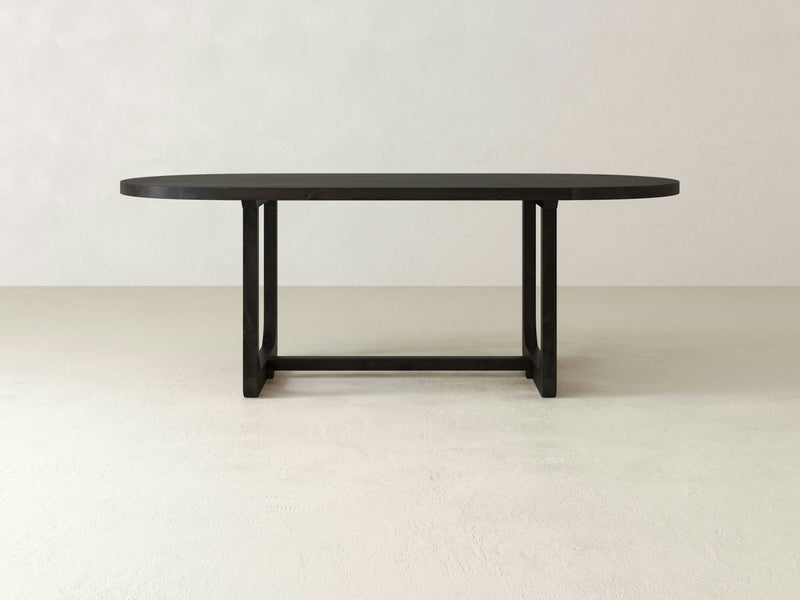 The Emersen Oval Dining Table - Charred Ember from James+James features a long, oval-shaped black surface with a simple, minimalist design. The table is supported by four legs connected by a rectangular support frame at the base and is set against a plain, light-colored background.