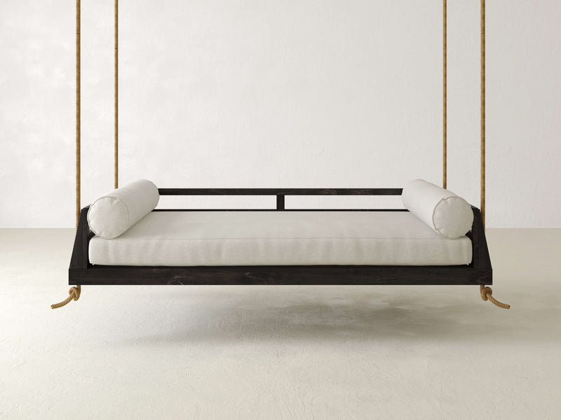 The Piper Porch Swing - Charred Ember by James+James features a minimalist wooden hanging daybed with a black frame, upholstered in white fabric, and suspended by four thick ropes. The frame maintains a minimalist design and includes two cylindrical cushions, one on each side. The background is a plain white wall and floor.