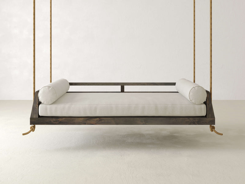 The Piper Porch Swing - Deep Grey by James+James features a wooden frame suspended by thick ropes in an empty white room. This minimalist swing bed is adorned with a light-colored mattress and two cylindrical bolster pillows on either side, creating a serene and uncluttered setting.