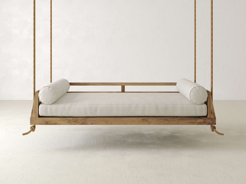 The Piper Porch Swing - Harvest Wheat by James+James, featuring beige cushions and two cylindrical bolster pillows, hangs from thick ropes in a bright, neutral-colored room. This minimalist wooden swing bed boasts a simple, natural design that creates a serene and inviting atmosphere.