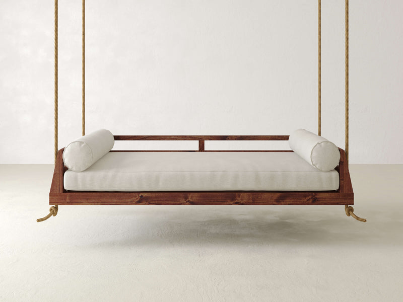 The Piper Porch Swing - Tuscany from James+James is a minimalist wooden daybed with a simple frame that hangs from ropes against a plain, light-colored wall. It features light-colored padding and comes with two cylindrical bolster pillows on either end.