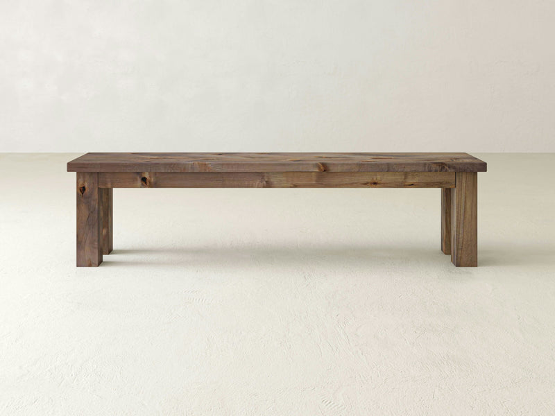 A minimalist Farmhouse Bench - Barn Wood by James+James is placed against a plain off-white background. The bench has a simple, rectangular design with four sturdy legs, showcasing the natural wood grain pattern.