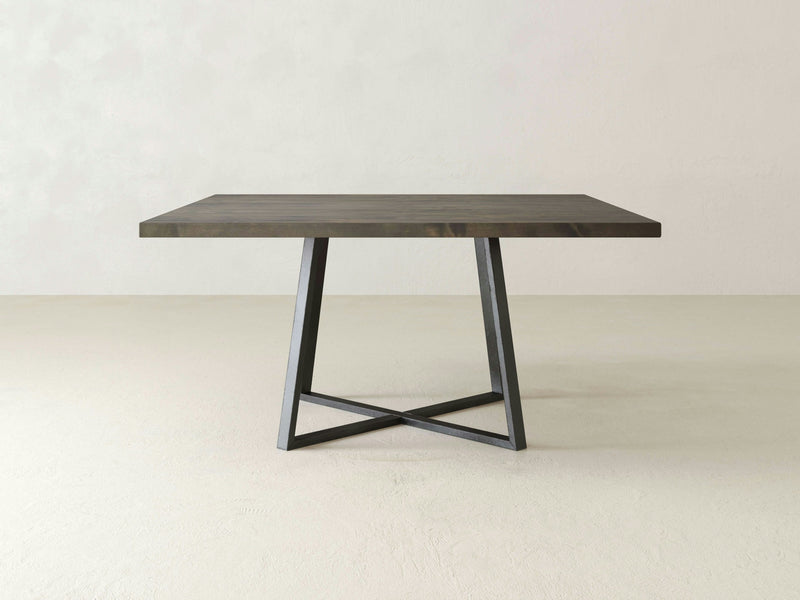 The Watson Square Dining Table by James+James features a modern, minimalist design with a deep grey wooden top and a black metal X-shaped base, all set against a plain, light-colored background.