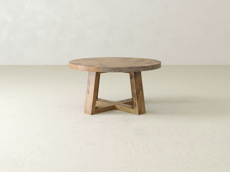 The Rae Coffee Table - Harvest Wheat by James+James features a round wooden design with a flat top and thick edges, standing on a cross-legged base. It boasts a natural, rustic finish and is set against a plain, light-colored backdrop in an empty room.