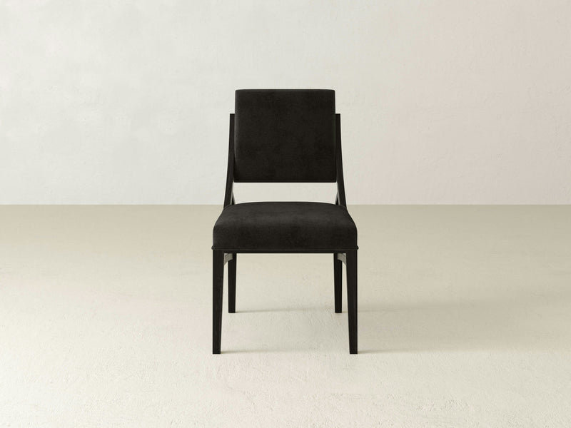 A Quinn Dining Chair - Charred Ember by James+James, featuring a sleek black design with a square backrest and slightly angled legs, is placed in the center of a plain room with light-colored walls and floor. Its minimalist style creates a stark contrast against the neutral background.