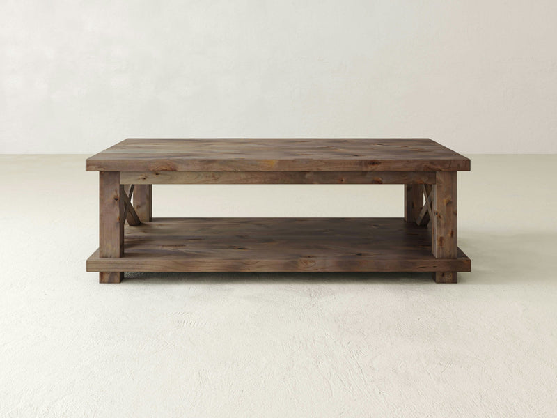 The Carter X Coffee Table - Barn Wood by James+James is a rectangular wooden coffee table that boasts a rustic finish. It includes a lower shelf for extra storage and decorative X-shaped side supports, offering both sturdiness and farmhouse-inspired design. The background is plain white.