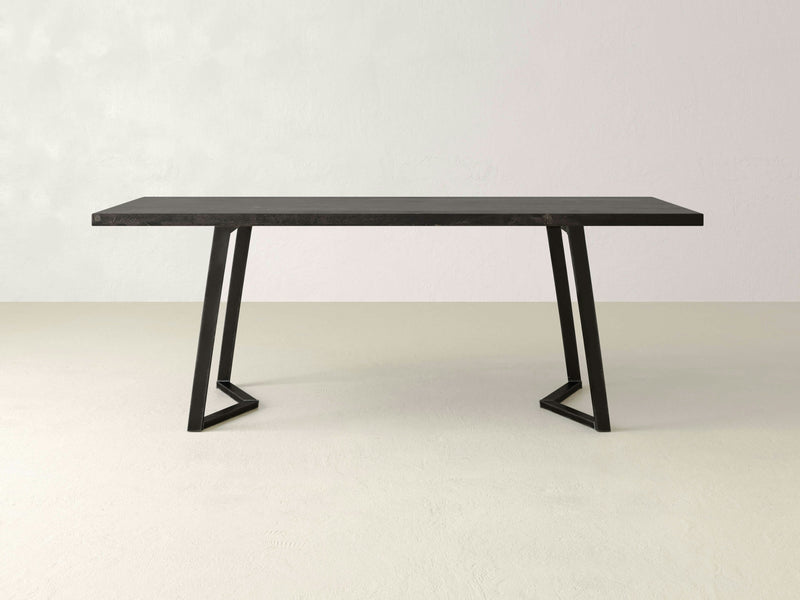 The Arkwright Dining Table - Charred Ember from James+James is a modern, minimalist rectangular table featuring a black top and angled metal legs. The smooth surface complements the sleek geometric design of its legs. The table is showcased against a plain, light-colored background.