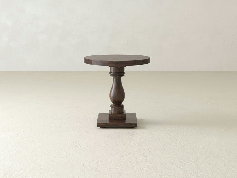 A compact, round wooden side table known as the Vivien Side Table - Tobacco by James+James stands alone in an empty room. It features a single ornate pedestal leg and a square base, finished with a polished surface. The background consists of plain, light-colored walls and a matching floor.