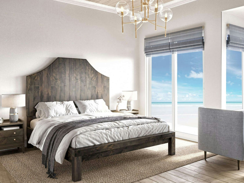 A modern bedroom features the Megan Bed by James+James in a deep grey finish, flanked by two nightstands with lamps. A large window offers a view of a sunny beach and ocean. A gray armchair is placed near the window, and the decor is minimalist with neutral tones.