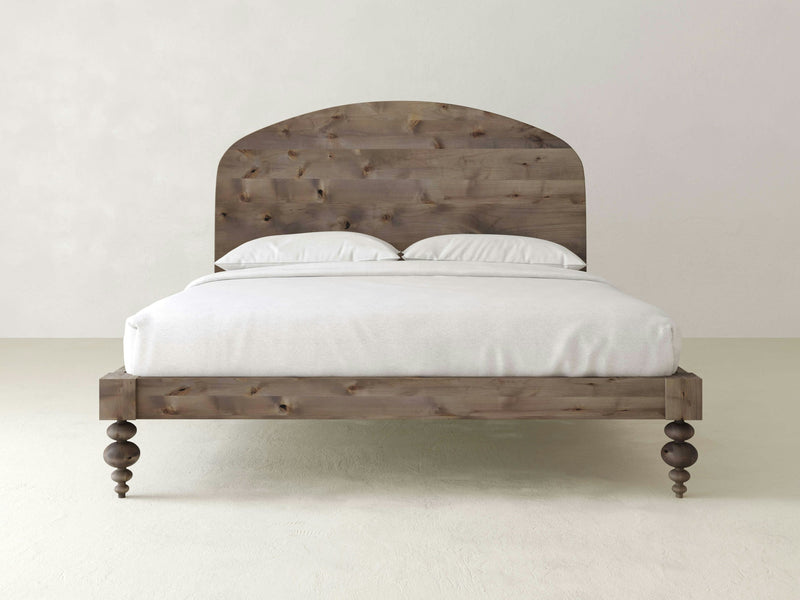 The Eden Bed - Barn Wood by James+James features a rustic wooden frame with a curved headboard. It is dressed in white bedding and includes two white pillows. The bed, which boasts uniquely turned wooden legs, is set against a plain off-white background.