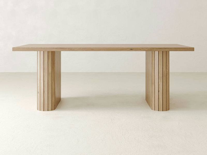 The Fern Dining Table by James+James is crafted from solid white oak hardwood and features a minimalist rectangular top with two thick cylindrical legs adorned with vertical grooves. Set in a sparse, light-colored dining room with a plain background and floor, it highlights the table's simple and modern design.