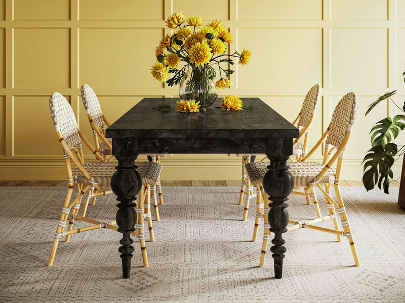 A dining area featuring the Bailey Dining Table in Charred Ember from James+James, accompanied by six wicker chairs, set against a yellow paneled wall. A glass vase with yellow flowers serves as a centerpiece on the table. The space is enhanced by a patterned gray rug and a potted plant in the corner.