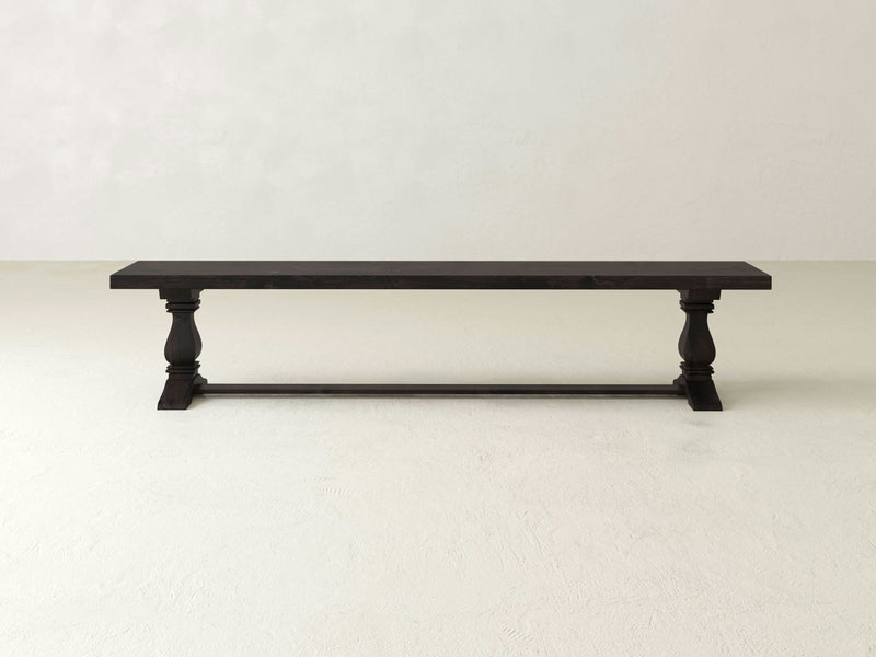 The Heirloom Bench - Charred Ember by James+James is a long, dark wooden bench with a simple, elegant design. This bench boasts a rectangular top and is supported by two sturdy, turned legs connected by a stretcher at the bottom. It is situated in an empty, light-colored room.