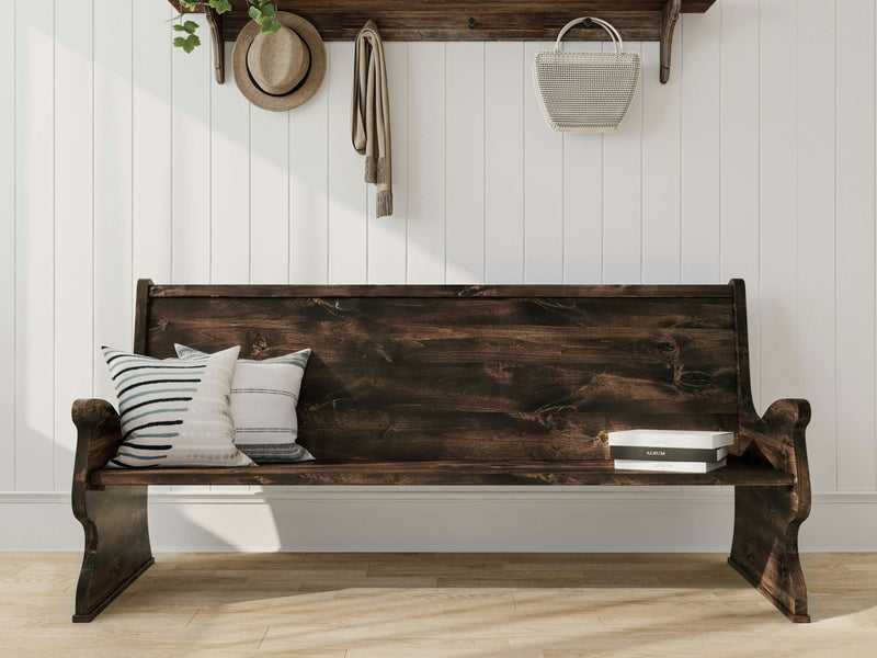 The Rustic Church Pew Bench - Tobacco from James+James is placed against a white shiplap wall. Resting on the bench are two pillows; one with stripes and another plain. Above it, a wooden shelf showcases a hat, a hanging plant, a handbag, and a scarf. Books are neatly positioned at the right end of the bench.