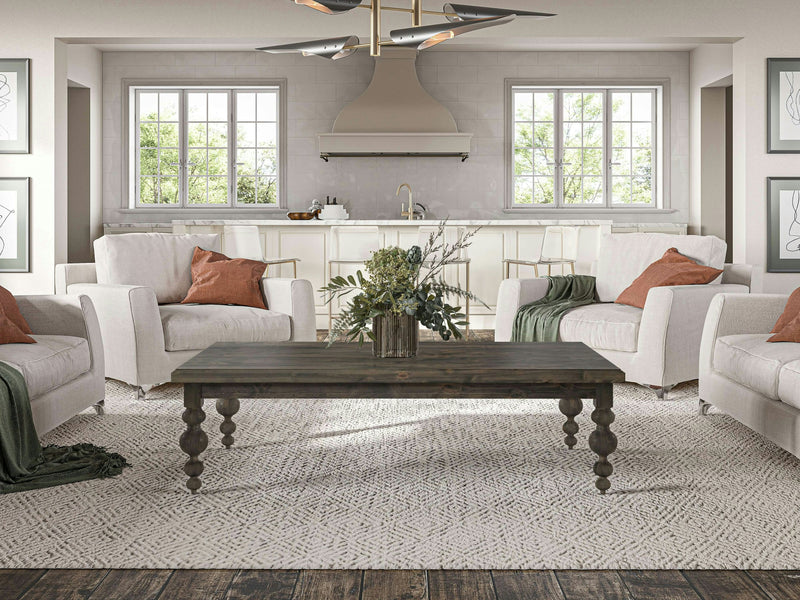 A cozy living room features four beige armchairs with rust-colored and green throw pillows, arranged around an Olivia Coffee Table in Deep Grey by James+James, adorned with a plant centerpiece. In the background, large windows bring in natural light, illuminating a modern kitchen space.