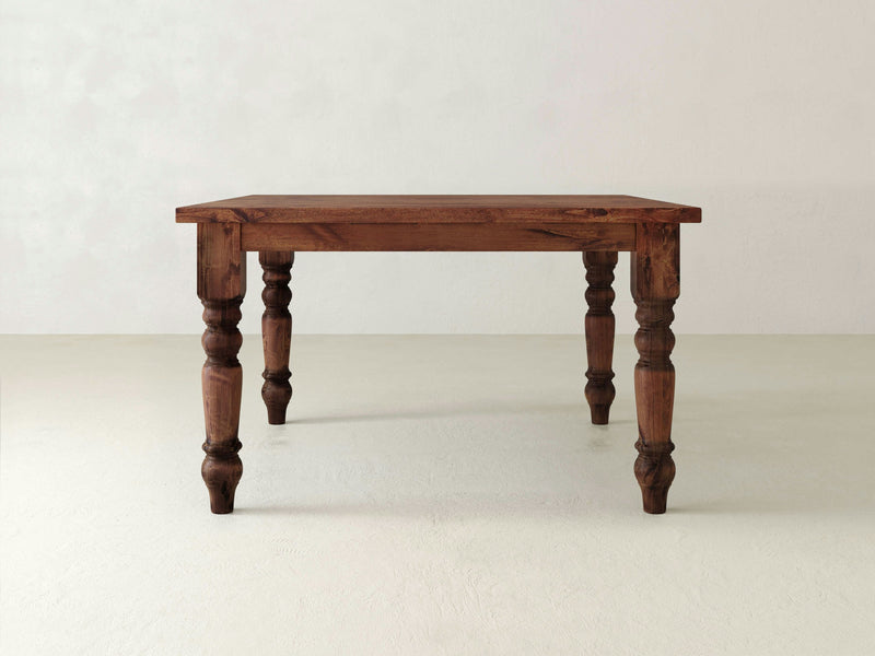 The Baluster Square Dining Table - Tuscany by James+James features a rectangular top and four intricately carved legs, set in a minimalist environment with a light-colored floor and a white wall backdrop. The table boasts a rustic look with its natural wood finish.