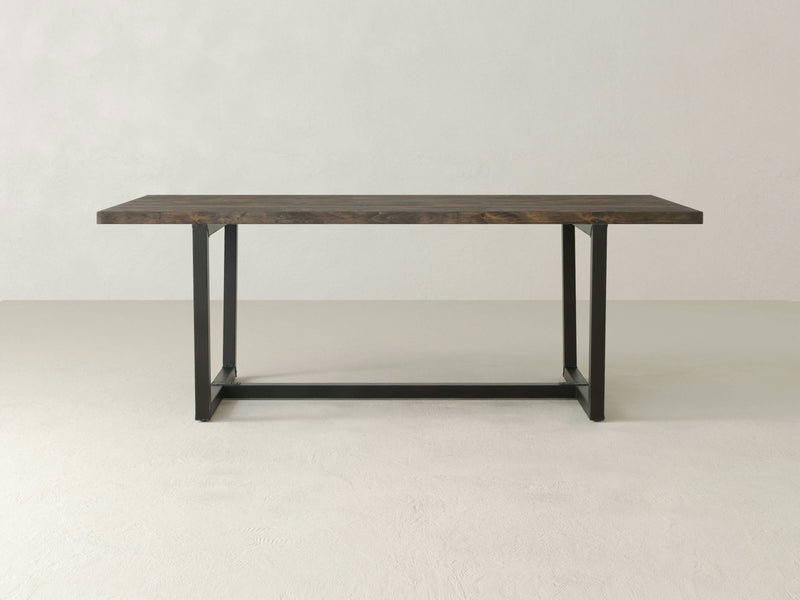 Introducing the Trapezoid Dining Table in Deep Grey by James+James. This minimalist wooden table features a dark brown top and black metal legs, crafted in a rectangular shape with a simple, modern design. It stands on a light-colored floor against a plain, light backdrop, showcasing its clean and sleek aesthetic perfectly.