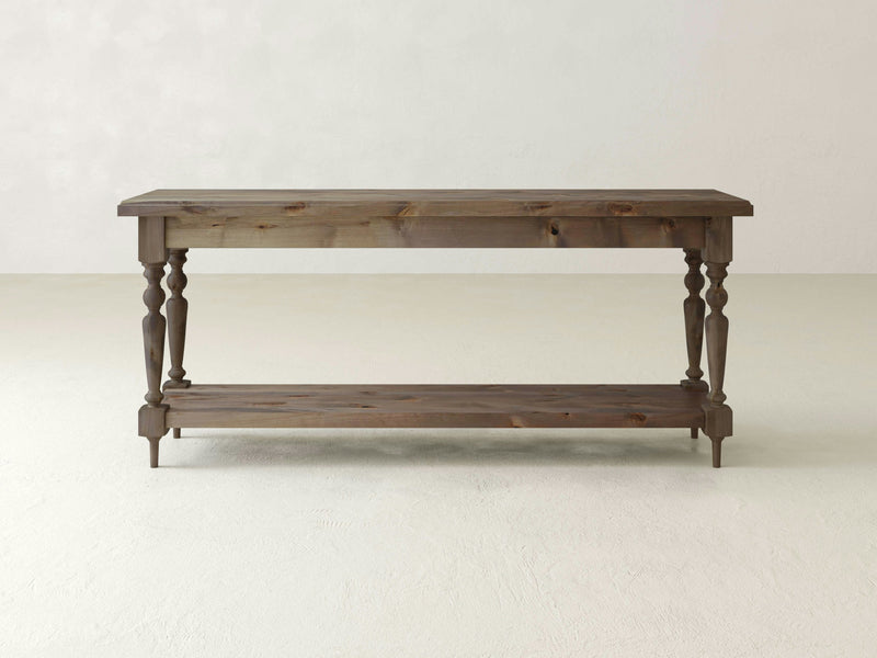 The Ella Sofa Table - Barn Wood by James+James is a rectangular wooden console table featuring four intricately carved legs connected by a lower shelf. It boasts a rustic, natural finish and is situated against a plain, light-colored wall on a light floor.