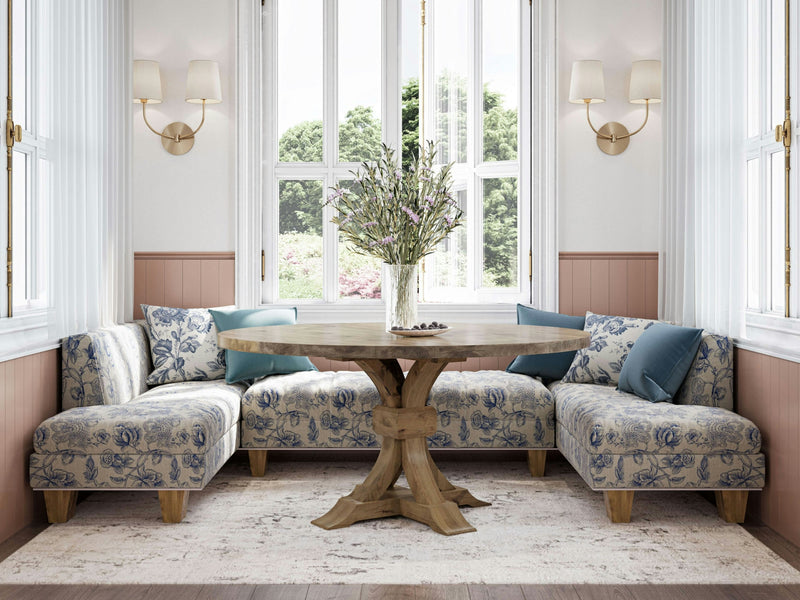 A cozy dining nook featuring an L-shaped cushioned bench adorned with blue floral upholstery, centered around the Violet Round Dining Table in Harvest Wheat by James+James. The scene is enlivened with a vase of flowers and illuminated by natural light streaming through large windows, complemented by two elegant wall sconces. Light-colored walls and a soft rug complete this inviting setting.