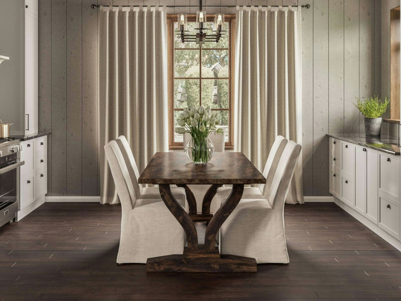 A cozy dining room featuring the Vera Expandable Dining Table in Tobacco by James+James, accompanied by six beige upholstered chairs. The table is adorned with a vase of white flowers. A large window draped with beige curtains is flanked by cream cabinets, while a dark wood floor enhances the earthy tones of the room.