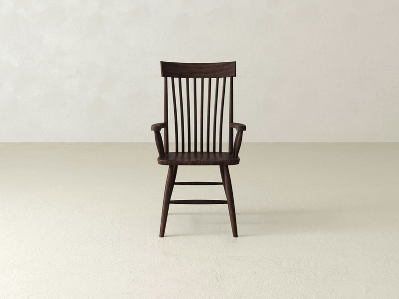 An Eleanor Arm Dining Chair - Tobacco by James+James, characterized by its wooden construction and dark brown finish, showcases a high back with vertical slats, armrests, and tapered legs. It is placed against an off-white wall and light-colored floor, embodying a minimalist and modern design.