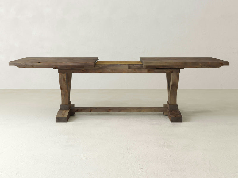 A close-up of the Vera Expandable Dining Table by James+James, showcasing the barn wood surface with a prominent natural knot. The wood grain is visible with wavy lines and variations in color from light to medium brown. A large crack extends from the knot, adding a rustic and textured appearance.