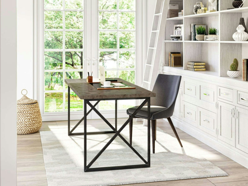 A well-lit home office with a large window features the Luca Desk in Barn Wood by James+James, which sits on a white rug. The desk is paired with a gray chair and holds a white lamp, notebooks, and various office supplies. Behind the desk stands a white built-in bookshelf adorned with decor items and green plants.