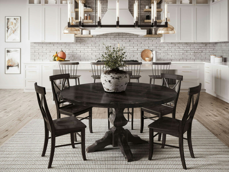 A modern dining room showcases the Sybil Round Dining Table in Charred Ember by James+James, accompanied by six matching chairs. A potted plant centerpiece adorns the table, while the background includes white cabinets, a brick backsplash, and two gold pendant lights above the kitchen island. The floor is accented with a large area rug.