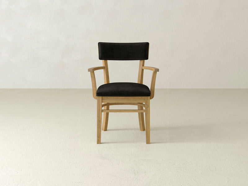 The Everly Arm Dining Chair by James+James, featuring Harvest Wheat wood and black cushions on both the seat and backrest, is placed against a plain, light-colored wall. The chair includes armrests and boasts a simple, minimalist design.